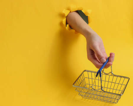 Minimalism shopping concept. Female hand holds a shopping basket through a torn hole in yellow backgroundの写真素材