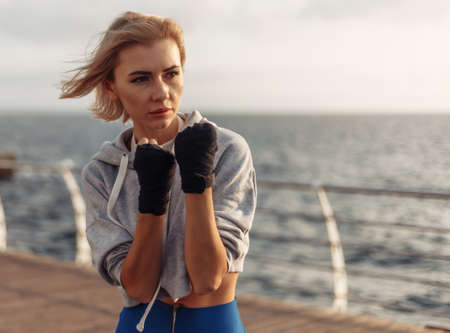 Young woman kickboxer in sportwear with wrapped hands in bandages trains on the beach at sunrise....