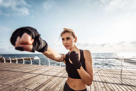 Young woman kickboxer with wrapped hands in bandages trains punch on the beach at sunrise. Morning training fighterの写真素材