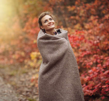 Young cheerful frozen woman wrapped in a plaid in the autumn forest with reddened leaves. Art portraitの写真素材