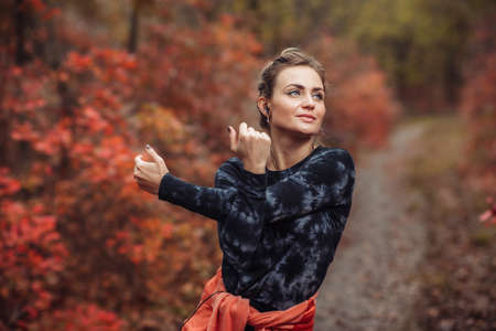Young attractive sport woman in sportswear doing stretching exercise for hand in autumn forest with reddened leaves of trees. Warm up before training. Healthy lifestyle conceptの写真素材