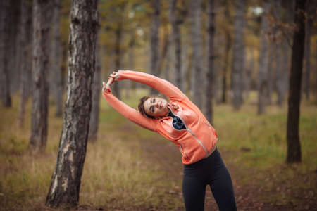 Young attractive sport woman in sportswear doing stretching exercise for hand in autumn forest. Warm...