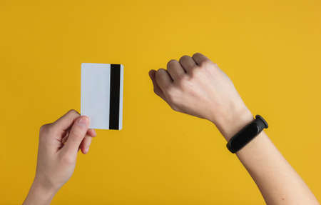 Hand with smart bracelet holding bank card on yellow background. Cashless payment, online shopping....