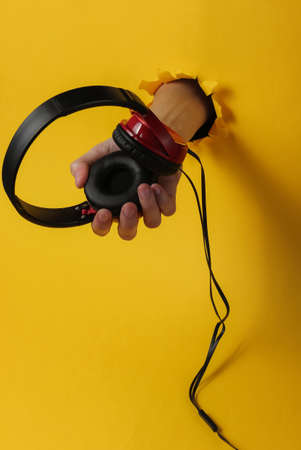 Female hand holds wired headphones through a torn hole in a yellow background. Minimalistic music...