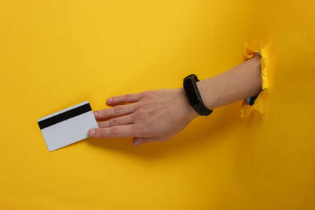 Hand with smart bracelet holding bank card on yellow background through torn hole. Cashless payment,...