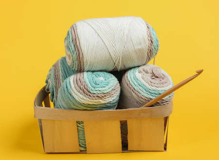 Skein of woolen threads of pastel colors and crochet hook in basket on yellow background. Housewives Hobbiesの写真素材