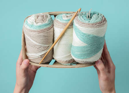 Women's hands holding skein of woolen threads of pastel colors and crochet hook in basket on blue...