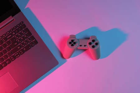 Gaming concept. Gaming equipment. Laptop with gamepad. Neon red-blue gradient light. Top viewの写真素材