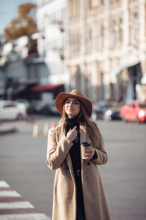 Young stylish woman dressed in a coat and felt hat walks around the city and drinks coffee on the go. Autumn timeの写真素材