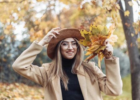 Young funny woman dressed in a coat and hat holds an armful of fallen leaves in the autumn park. Autumn funの写真素材