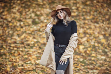 Image-shot of a young attractive woman wearing a coat and felt hat on blurry background of fallen leaves in the autumn parkの写真素材