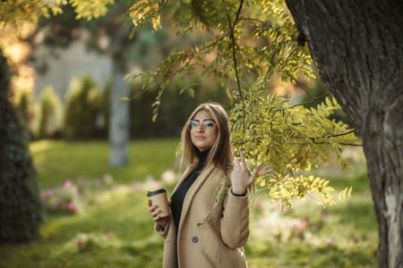 Young attractive woman dressed in stylish coat, turtleneck posing in the autumn park and holds cup...