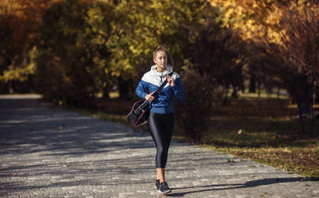 Fit slim woman in sportwear and a training bag on her shoulder walks through the autumn park at trainingの写真素材