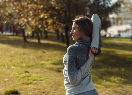 Young sport woman in a sports suit is warming up in the autumn park. Stretching hands before training. Fitness training outdoorsの写真素材