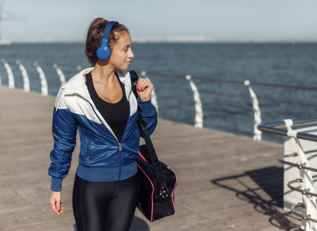 Young fit woman with a training bag and headphones walking along the beach promenade on a bright sunny dayの写真素材
