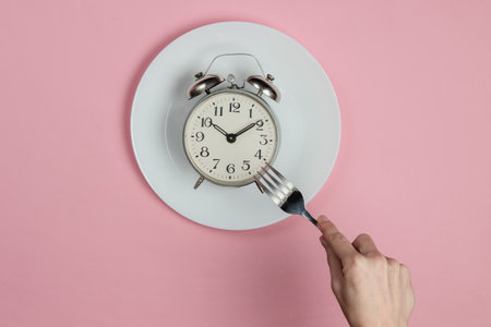 Breakfast time! Female hand with fork about to eat from a plate with a retro alarm clock on pink...