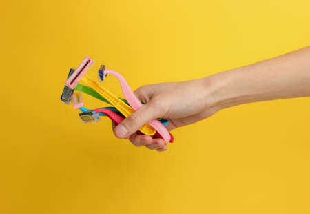 Female hand holds lot of colored disposable razors of epilators on yellow background. Beauty and fashion shotの写真素材