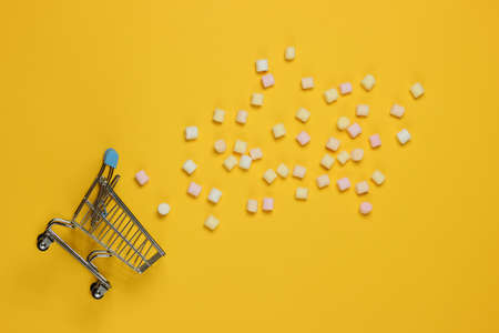 Lot of mini marshmallows and shopping trolley on yellow background. Top view