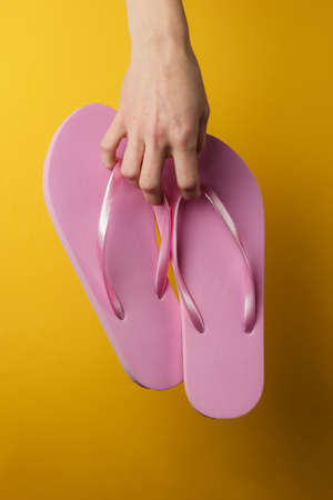 Studio fashion shot. Female hands holding pink flip flops on a yellow background. Beach vacation. Summer timeの写真素材