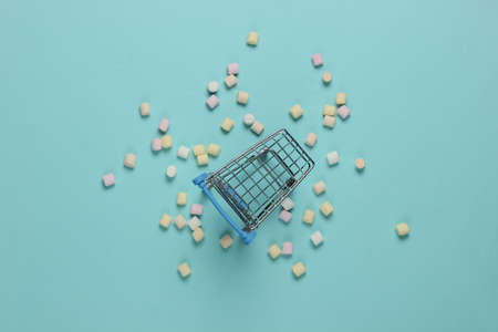 Lot of mini marshmallows and shopping trolley on a blue background. Pastel color trendの写真素材