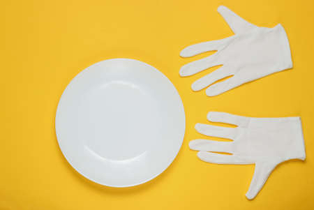 White waiter gloves and empty ceramic plate on yellow studio background. Top viewの写真素材