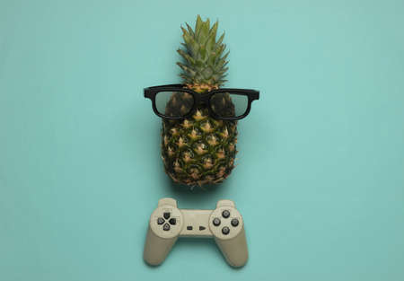 Gamer concept. Fun and humor. Pineapple in sunglasses and gamepad on blue background. Top view