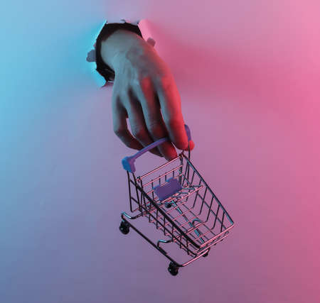 Hand holding shopping trolley through torn hole. Creative pop art pink blue neon color. Trendy...