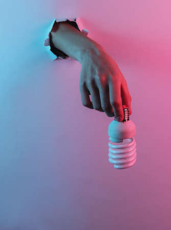Hand holding eco spiral light bulb through torn hole. Creative pop art pink blue neon color. Trendy gradient illumination. Night lightの写真素材