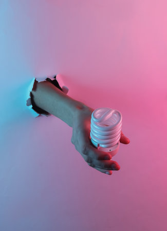 Hand holding eco spiral light bulb through torn hole. Creative pop art pink blue neon color. Trendy...