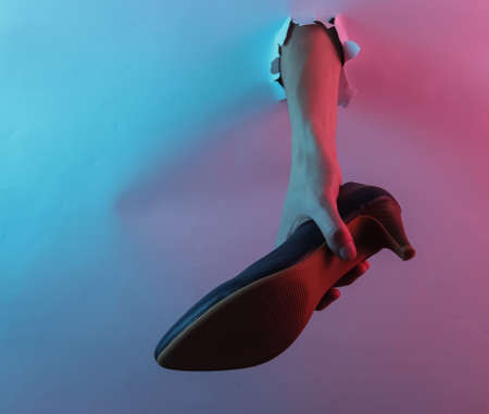 Hand holding high heel shoes through torn hole. Creative pop art pink blue neon color. Trendy...