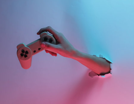 Hand holds gamepad through torn hole. Creative pop art pink blue neon color. Trendy gradient illumination. Night lightの写真素材