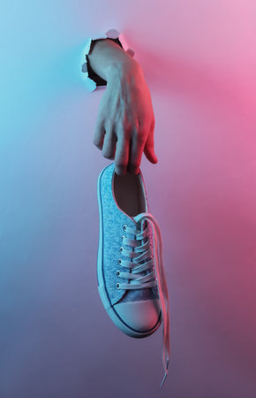 Hand holding sneaker through torn hole. Creative pop art pink blue neon color. Trendy gradient illumination. Night lightの写真素材