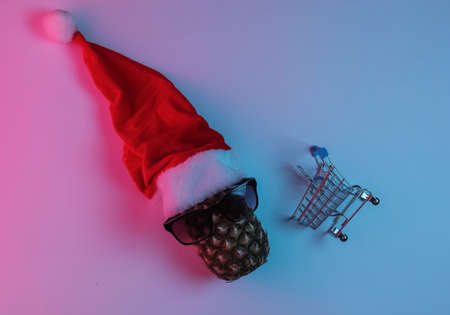 Christmas shopping. Pineapple dressed in Santa hat with sunglasses and shopping trolley. Neon Blue Pink Night Light. Top viewの写真素材