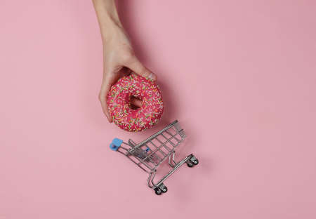 Minimalistic food still life. Hand holding Glazed donut and shopping trolley on a pink pastel...