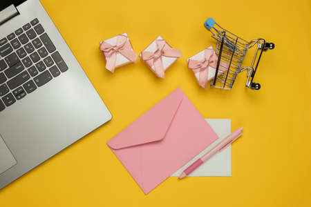 Laptop, envelope with letter and pen, boxes of gifts and shopping trolley on yellow background....