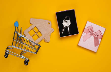 Keys in a gift box with a bow, house figure, shopping trolley on a yellow background. Top viewの写真素材