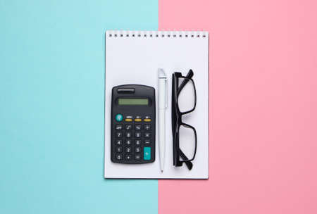Notebook with pen, calculator and glasses on pink blue pastel background. Top viewの写真素材
