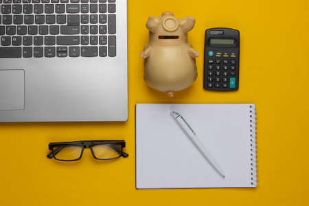 Calculation of the family budget. Calculator, laptop, piggy bank, notepad on a yellow background. Top viewの写真素材