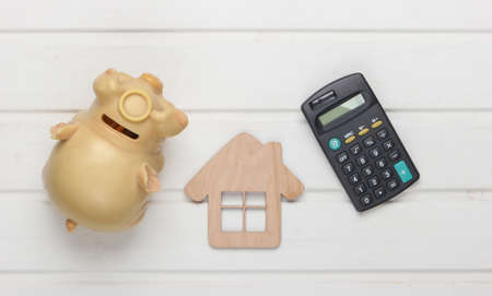 Calculation of rent payment for housing. Creative flat lay composition. House figurine, calculator, piggy bank on a white table. Top viewの写真素材