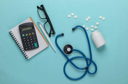 flat lay medical composition. Economic analysis. Calculator with notepad, stethoscope and pills on a blue pastel background. Top viewの写真素材
