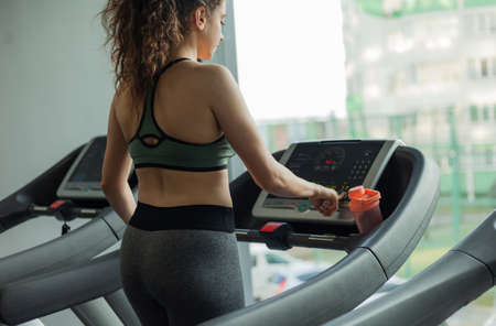 Fit woman in sportswear runs on a treadmill and looks at her watch in the gym. The concept of a healthy lifestyle, warming up, fitness, weight loss.の写真素材