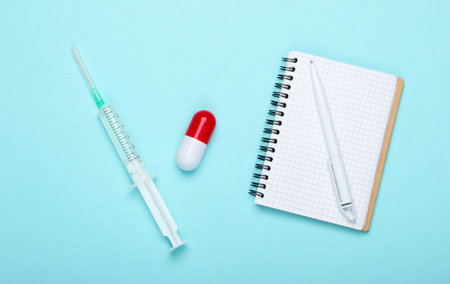 Flat lay medicine composition. Pill capsule, syringe with vaccine, notebook on a blue pastel background. Top viewの写真素材
