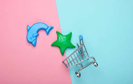 Shopping trolley with toy forms for the sand (beach toys) on a blue-pink pastel background. Top view