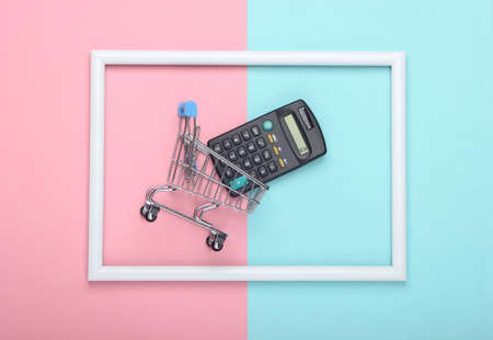 Shopping trolley with calculator in a white frame on pink blue pastel background. Top view. Flat layの写真素材