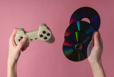 Hands hold cd discs and gamepad on pink pastel background. Gaming, recreation and entertainmentの写真素材