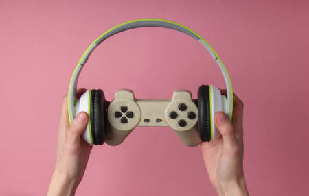Hands hold stereo headphones and gamepad on pink pastel background. Gaming, recreation and entertainmentの写真素材