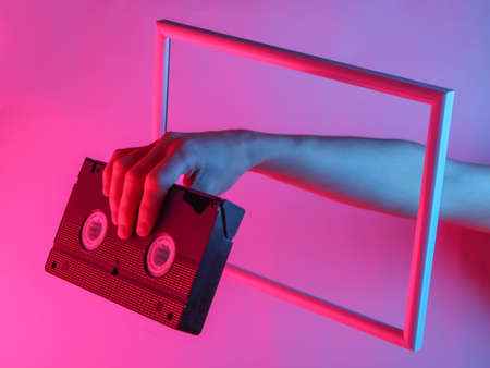 Creative art. Female hand holding video cassette through a soaring frame with neon holographic light. Gradient glow.の写真素材