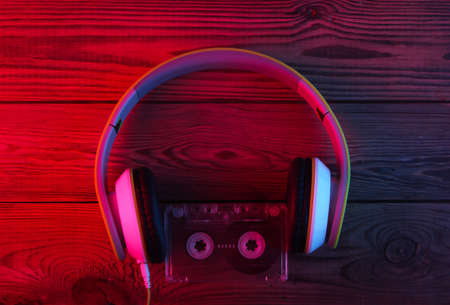 Stereo headphones with audio cassette on wooden background. Neon red blue light. Gradient glow....