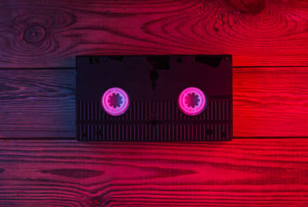 Video Cassette on wooden background. Neon red blue light. Gradient glow. Retro media and...