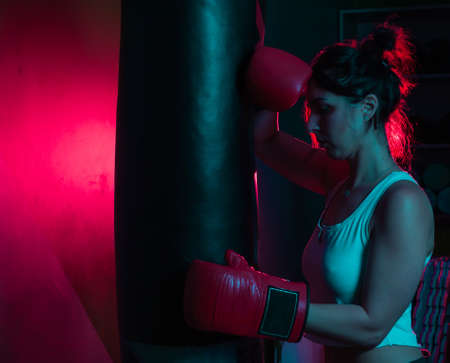 Tired woman boxer in boxing gloves is leaning on a punching bag in red blue neon light over dark...